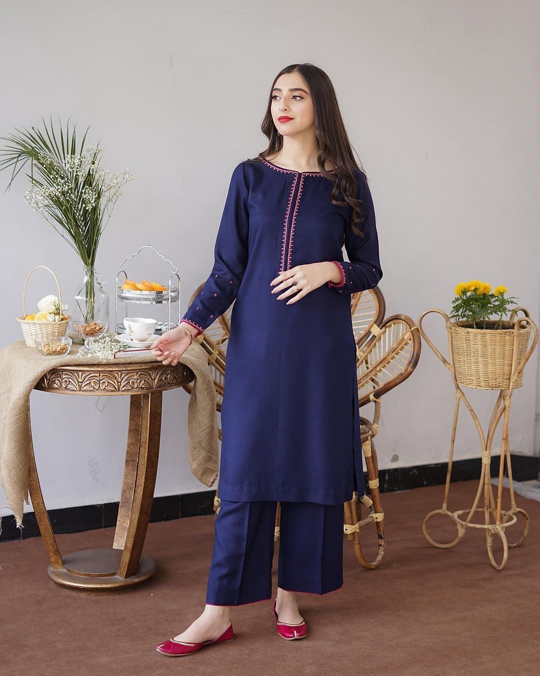 Woman in a blue outfit standing in a room with a table and decorative items.Elegant Pakistani winter 3pc suit in linen fabric